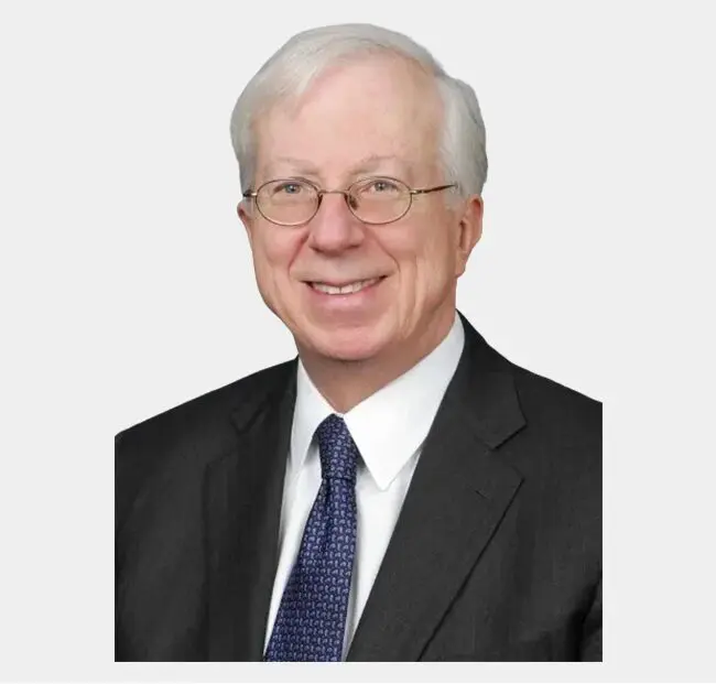 Portrait of an elderly caucasian man with glasses, smiling, wearing a dark suit and a light blue tie against a gray background.